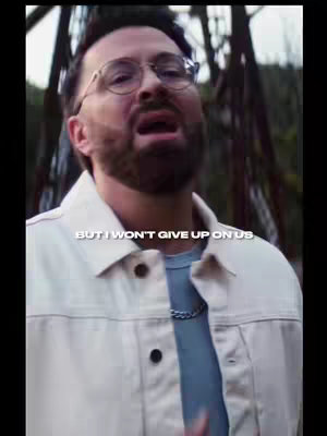 The cover image shows a man with a beard, glasses, and a light-colored jacket, appearing to speak into a camera. In the background, there's a blurred, metallic structure.