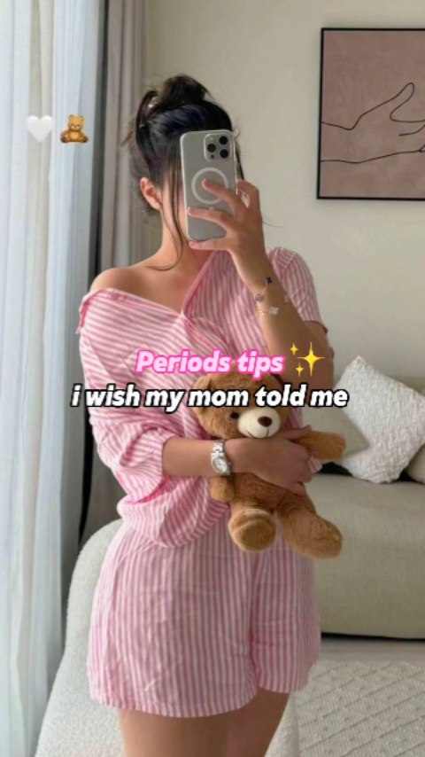 The image shows a woman in a pink striped pajama set holding a teddy bear. She is taking a selfie with her phone, with the camera obscuring her face. The background shows a room with a couch, pillows, and a piece of art on the wall.