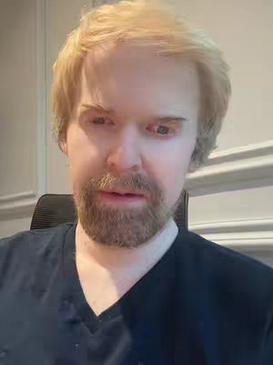 A man with light hair and a beard looks directly at the camera. He is wearing a dark v-neck shirt and is seated in front of a wall. The background includes a white wall with decorative panels. The overall composition is a close-up shot focusing on the man's face.