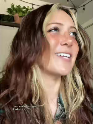 A woman with wavy brown and blonde hair is seen smiling widely and looking up and to the side. There is a close-up of her face, showing a joyful expression. Text overlayed on the cover image: 'you actually brought loaded dice.'