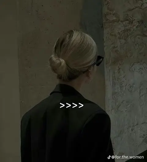 The image shows a woman seen from the back, wearing a black blazer. Her blonde hair is styled in a low bun. The background appears to be a textured wall, with the composition emphasizing the woman's hairstyle and attire. Below the woman's back, a series of greater-than symbols are used as an added element.