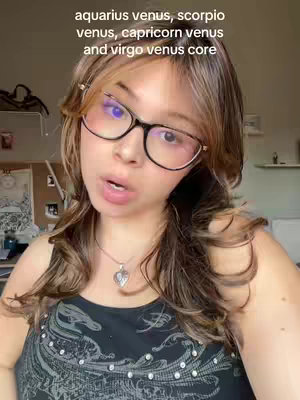 A young woman with fair skin and medium-length, dark-blonde hair is seen in a selfie-style video. She wears large, round-framed glasses and a black tank top. Text overlays at the top read 'aquarius venus, scorpio venus, capricorn venus and virgo venus core'. She is facing directy to the camera with her mouth slightly open. The background appears to be an interior space with a glimpse of photos and documents on the wall.