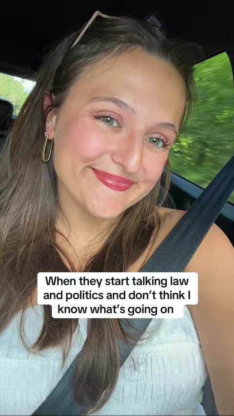 A selfie-style shot of a young woman seated inside a car, likely recording a video. She is smiling and the background outside the car window is blurred green. The woman has long brown hair, a light complexion, and is wearing an open-necked white top. The text overlay in white font reads, 'When they start talking law and politics and don't think I know what's going on'.