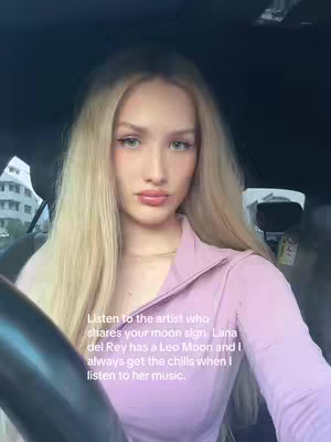 A young woman with long blonde hair gazes directly at the camera while seated inside a car. The background is a blurred mix of interior car elements and an exterior view, suggesting a daytime setting. The image is well-lit, with the woman as the primary focus.