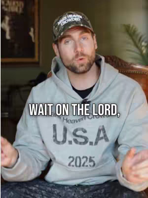 A man in a gray hoodie and a camouflage baseball cap is the central focus. The text overlay reads, 'WAIT ON THE LORD.' The background appears to be an interior setting with a painting and a glimpse of greenery on the right. The composition highlights the man and the message.