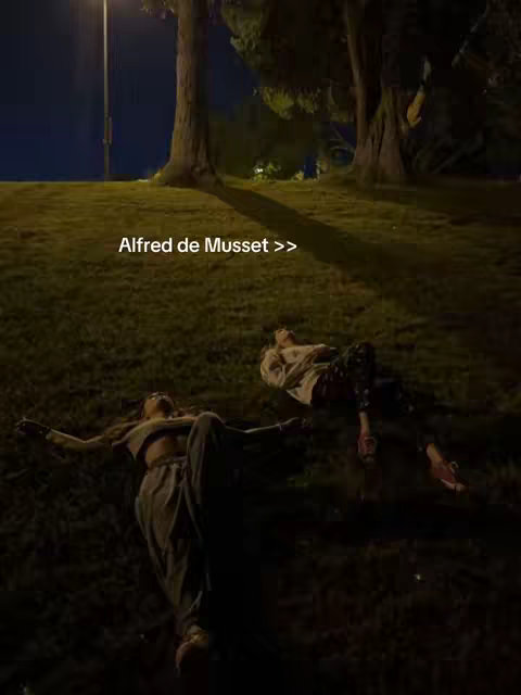 Two people are lying on a grassy hill at night. The sky is dark, and there is a bright streetlight in the background.