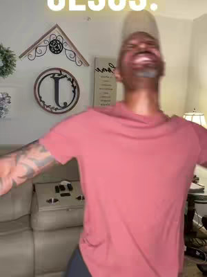 The image features a man in a pink shirt with his arms outstretched, giving a joyful expression. He seems to be inside a house with a wall in the background and some decorations, including a wooden sign, indicating a possible home setting. The shot is slightly blurred, potentially indicating movement or a focus on the man's expression. The word "JESUS" is written above the man