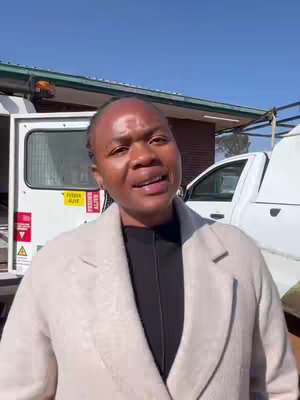A woman stands outdoors, facing the camera. She is wearing a light-colored coat over a black top. In the background, there is a white vehicle and a building. The woman is speaking.