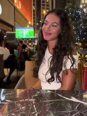 The cover image shows a woman seated at a table in an outdoor setting, likely a restaurant or bar. The background includes blurred elements of city buildings and lights, suggesting night-time. The woman is the focus, wearing a white off-the-shoulder top, her dark hair long and wavy. She smiles, creating a relaxed and inviting expression. She appears to be in an intimate moment.