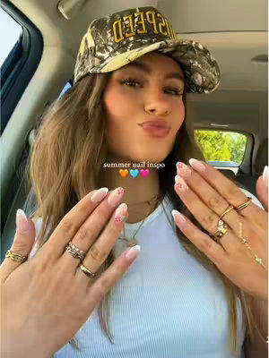 A selfie taken from inside a car featuring a young woman. She is wearing a camouflage baseball cap and a white t-shirt. Her hands are up in front of her, adorned with multiple rings and manicured nails with a white French tip and floral design. The hook 'summer nail inspo' is in the middle of her face.