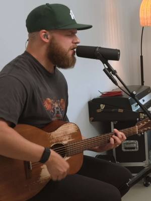 A man plays an acoustic guitar and sings into a microphone. He's wearing a baseball cap and a dark t-shirt. The setting appears to be a studio with simple, off-white walls and a lamp in the background.