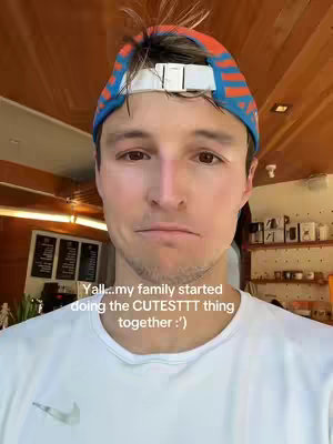 A selfie-style shot of a young man in a white t-shirt and a blue and orange cap. He appears to be indoors with a wooden background, he has a neutral expression. Text overlays at the bottom.