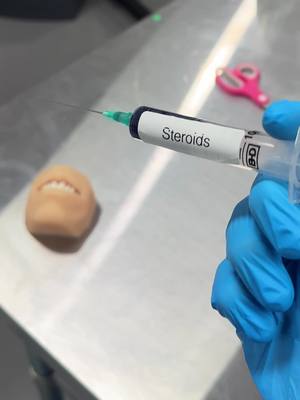 The image features a close-up shot of a medical or laboratory setting. A hand wearing a blue glove holds a syringe filled with a clear liquid. The syringe is labeled with the word "Steroids." In the background, out of focus, are a small plastic human face replica with visible teeth and a pair of pink scissors. The overall aesthetic has a clinical, possibly humorous, tone.