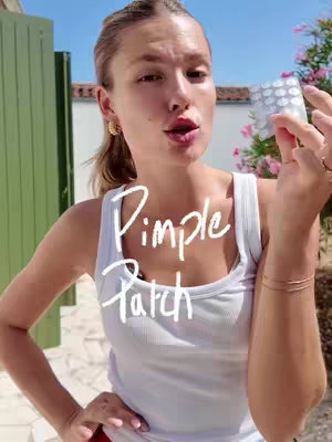 A woman wearing a white tank top stands outdoors, holding pimple patches. Text overlayed on image reads 'Pimple Patch'. The background shows a blurred green gate, blue sky, and foliage.