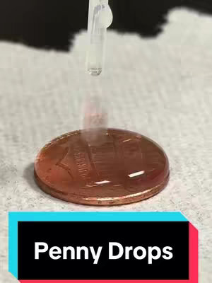 The image shows a close-up view of a penny coin with a drop of a liquid placed on top of it, held there by surface tension. A clear tube with a tip is visible dropping the liquid. The text overlay at the bottom says 'Penny Drops'. The background is blurred, suggesting a lab or experiment setting.