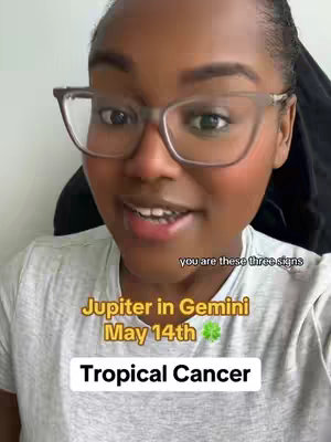 The image is a selfie-style shot featuring a woman wearing glasses, looking directly at the camera and speaking. Text overlays include astrological references, such as "Jupiter in Gemini," "May 14th," and "Tropical Cancer," along with a small green shamrock symbol. The woman's face is well-lit, showing her expression in a medium close-up. The overall composition utilizes vibrant colors and clear text for an informative and engaging visual.