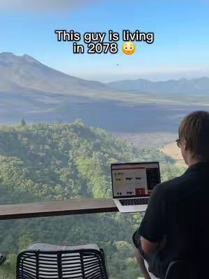 A man sits with his back to the camera, working on a laptop at a table that seems to be on a scenic overlook. Ahead of him, there's a vast landscape of rolling hills, trees, and a large mountain range under a clear blue sky. Text overlayed on the image states, 'This guy is living in 2078.'