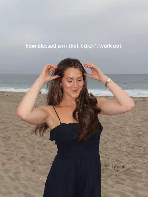 A woman in a navy dress stands on a sandy beach with the ocean and a cloudy sky in the background. She is smiling and has her hands near her hair.