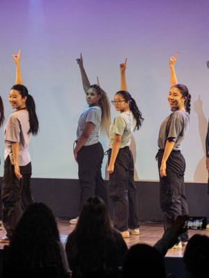 A group of young women are on a stage, lit by cool-toned lighting, with a stage backdrop and audience visible at the bottom. The women are dressed in basic grey t-shirts, black pants, and sneakers. They each have one arm raised, pointing upwards, with a slight lean, and the overall vibe suggests a dance performance. The audience can be seen in the lower part of the image.