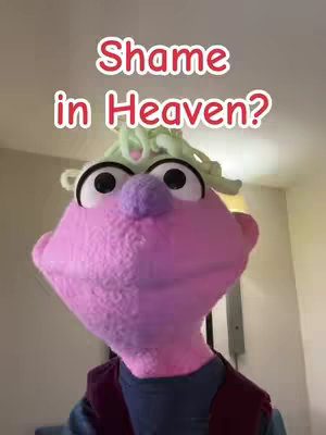 A close-up of a puppet with a pink face, large black eyes, a purple nose, and green hair. The words "Shame in Heaven?" are written in white, outlined in red, above the puppet's head.