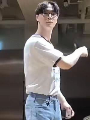 The image shows a man standing in a room, looking up and slightly to the left. He is wearing glasses, a white t-shirt with a black collar, and blue jeans. His right arm is extended to the side, and the background appears to be a simple, brightly lit wall. The overall composition has a casual, yet slightly posed feel.
