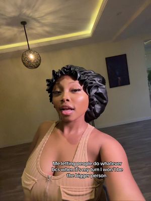 A woman with dark skin and a black bonnet is in what appears to be a well-lit interior. She is wearing a light-colored tank top, and her expression suggests she is speaking. The background is simple, with neutral-colored walls, a painting, and a patterned ceiling fixture.