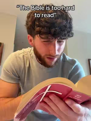 A man with curly brown hair and a beard is holding and looking into a Bible. He is wearing a green t-shirt and the background is out of focus suggesting an indoor setting. The book is open, revealing red and white pages and a prominent white cross design.
