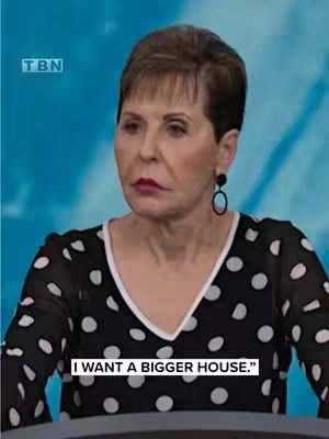The image features a medium shot of a woman, Joyce Meyer, against a blurred blue background. She has short, dark hair and is wearing a black and white polka-dotted top. The text overlay reads, "I WANT A BIGGER HOUSE." at the bottom of the image. A TBN logo is visible in the top left corner. The lighting appears to be studio-style, and the overall composition focuses on the upper body and facial expression of the woman.