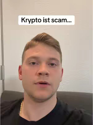 A man is centered in the image, facing the camera in a selfie-style shot. Above his head, a white rectangle with black text reads "Krypto ist scam...". He has short, light brown hair and is wearing a black shirt with a gold chain. The background appears to be a plain, light wall.