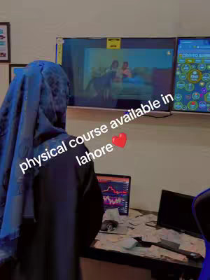 A person wearing a blue hijab is seen from the back, partially obscuring their face. In the background, there are two screens displaying content, a desk with a laptop showing a trading chart, and additional screens. Overlayed text reads 'physical course available in lahore' with a heart emoji.