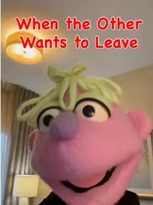 The video features a pink puppet with a green wig in the foreground. The puppet has large black eyes, a purple nose, and a slightly open mouth, creating a smiling expression. Above the puppet, the text overlay reads "When the Other Wants to Leave" in a red, stylized font. The background is slightly out of focus but includes a warm-toned lamp and a glimpse of a room.