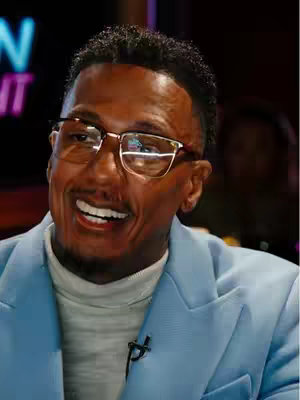 Nick Cannon smiling, wearing glasses and a blue blazer with a white turtleneck.