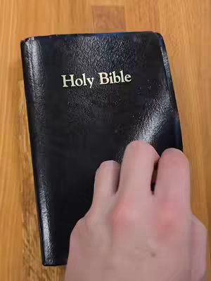 A tightly framed shot features a closed black book, likely a Bible, with the title 'Holy Bible' in gold lettering. A hand, with visible fingers and the side of the palm, is in the foreground, touching and slightly obscuring the book. The book rests on a light-brown, wood-grain surface, suggesting a table or desk.