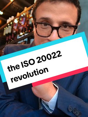 A man in glasses and a suit is partially visible behind a banner with text. The banner is primarily white with aqua on the top and crimson on the bottom. The text in the banner reads, 'the ISO 20022 revolution'. The background is blurred, appearing to be an interior space with lights.