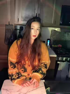 A woman with long brown hair and red lipstick poses in a kitchen. She is wearing a yellow patterned cardigan. The background includes white cabinets, a stove, and appliances.