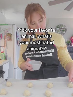 A woman with red hair is in a kitchen, holding a small bowl and placing cookie dough balls on a tray. Text overlayed on the top of the image reads: f*ck your favourite animal, what's your most hated animal??