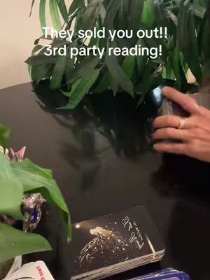 A dimly lit scene features a dark surface in the foreground, likely a table or desk. On the left, a lush green plant sits in a decorative pot. In the center, a deck of cards is held by a person's hand, with fingers adorned with a ring, preparing to deal. On the table, a partially visible book with golden lettering and a silhouette design rests in front of the cards . The background shows more foliage, indicating an indoor setting with a focus on spiritual or tarot-related content, as suggested by the text overlay. Overall, the image has a mystical and intimate feeling.