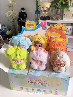 The cover image showcases an unboxing, with a box of 'Emotional Hairy Monsters' blindbox keychains prominently displayed. Six colorful plush toys with unique designs and expressions are visible. The background includes shelves with decorative items and figurines, suggesting a cute and organized setup.