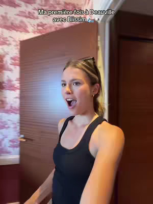 A selfie-style video cover image of a young woman with her mouth wide open in surprise. She is standing in a doorway, with a patterned wallpaper visible behind her, and a wooden doorframe to her right. Text overlayed on the cover image says, "Ma première fois à Deauville avec Blissim 🐳".