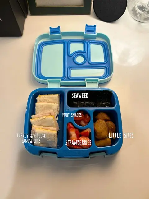 An overhead shot of a school lunch box open, displaying contents. The box is blue and light blue with compartments filled with food labeled with white text: "TURKEY & CHEESE SANDWICHES", "SEAWEED", "FRUIT SNACKS", "STRAWBERRIES", "LITTLE BITES".