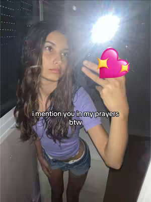 A selfie-style shot of a young woman with long brown hair, looking at the camera in a bathroom. She is wearing a light purple top and jean shorts. Her right hand is raised, displaying a pink heart sticker with yellow stars near her fingertips, with two fingers raised in peace sign. The background is a white tile. There's text overlaid that says, "i mention you in my prayers btw."