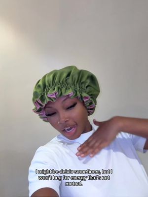 A selfie-style shot featuring a young, Black woman wearing a green and pink bonnet. She is looking slightly off-camera to the left with a soft smile, one hand gesturing towards her chest, and her lips parted. The background is a neutral, light-colored wall.