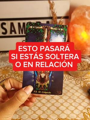A hand holding a tarot card, with a red banner displaying text hovering over the card. The background features a lit object, and part of a sign.