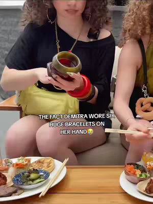 Two women seated at a table with food, one holding many bracelets, text overlay at the bottom. The image shows two women seated at a table with food in front of them. The woman on the left is holding a stack of colorful bracelets on one hand. Text overlays the image. The food on the table includes sushi and other dishes. The setting appears to be outdoors with some greenery visible.