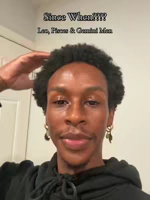 A selfie-style shot featuring a young Black man with dark, curly hair. He is wearing a black hoodie and small earrings. The man has a neutral expression, and the background is a plain wall with a white door. Text overlays the image.