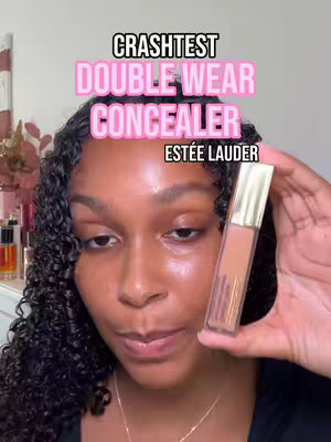 A woman with dark curly hair faces the camera, holding a makeup product. Text overlays read "CRASHTEST DOUBLE WEAR CONCEALER" and "ESTÉE LAUDER". The background includes glimpses of makeup products.