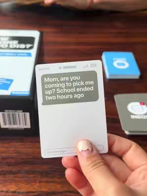A hand holds a card displaying a message conversation on a phone interface. The card reads, "Mom, are you coming to pick me up? School ended two hours ago." The setting includes other cards and is on a wooden surface.