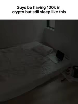 The image shows a dimly lit bedroom with a bed as the central focus. The bed is covered with a white sheet and a pillow, and a laptop is placed on the bed, suggesting a workspace or entertainment setup. The room is minimalistic, with a window visible in the background, creating a sense of isolation and quietness. The overall composition is simple and somewhat moody. The text overlayed says "Guys be having 100k in crypto but still sleep like this."