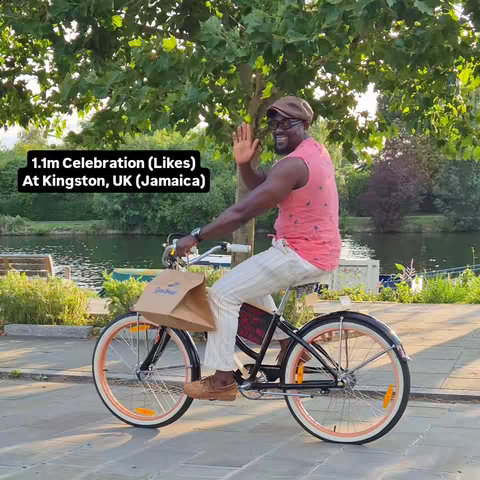 A man with a hat and sunglasses rides a bicycle with a paper bag on the front. He is waving with his right hand. Behind him is a lake with trees, and the sky is a light blue. The bike is black with orange tires.