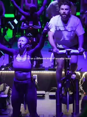 The cover image shows a fitness studio setup with several stationary bikes. A woman in athletic attire is riding a bike with her arms raised in the air, with a microphone near her mouth. Beside her, a man is riding another bike. They're in front of a group of other people on bikes. The lighting has a purple hue.